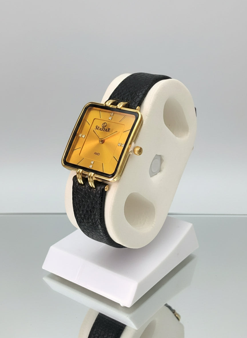 Gold rectangular watch on a black strap with a white display stand.